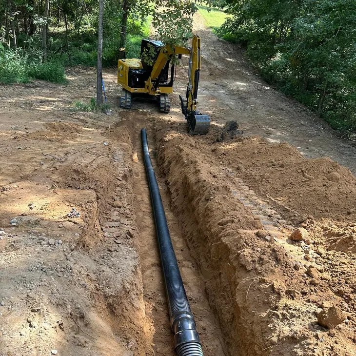 Corrugated drainage pipe set in an open trench alongside a gravel drive, with a compact excavator working the trench line on a Modern Shovel job